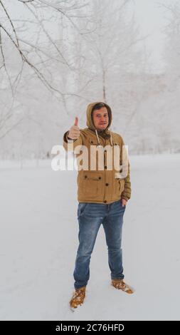 Schöner Mann, der im Winter im Park die Daumen hoch zeigt. Erwachsene junge Mann trägt beige Kapuzenjacke zeigt Geste der Zustimmung in Stockfoto