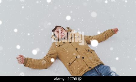 Porträt eines jungen Mannes in Jacke und Jeans, im Schnee liegend. Schöner Mann lügt, spreizt seine Hände und schließt seine Augen vor Freude im Winterpark Stockfoto