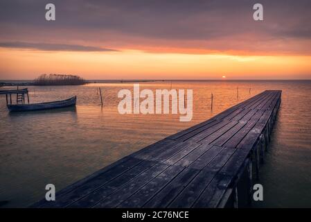 Sonnenaufgang oder Sonnenuntergang blick auf einen See an einem bewölkten windigen Tag mit einem hölzernen Ponton im Vordergrund und einem Fischerboot Stockfoto