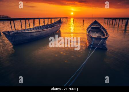 Zwei hinterleuchtete Fischerboote mit der Sonne im Hintergrund bei Sonnenaufgang oder Sonnenuntergang mit einigen Wolken am Himmel und zwei hölzernen Pontons Stockfoto