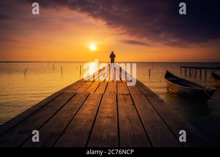 Einsame Person, die auf einem Ponton steht, meditiert und den Sonnenaufgang oder Sonnenuntergang auf einem See mit einem Fischerboot auf dem See genießt Stockfoto