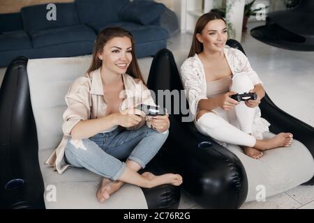 Lächelnde Freundinnen spielen Videospiele zu Hause, mit Joysticks und Spaß haben Stockfoto