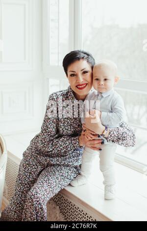 Porträt einer Großmutter, die auf einer Fensterbank sitzt und ihren Enkel umarmt Stockfoto