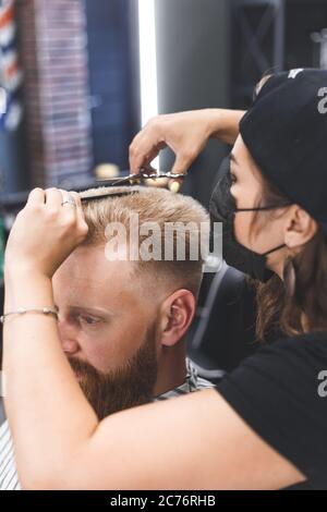 Frauen Friseur macht flache top Frisur mit Schere einen bärtigen Mann. Dienst in einer Maske von Virus. Stockfoto