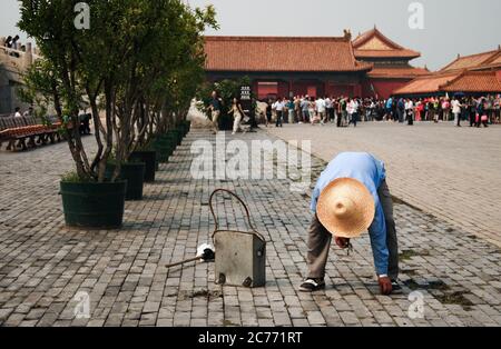 Ein älterer Hausmann, der den Müll aus dem Bereich der Verbotenen Stadt aufsammelt, trägt einen Strohhut und ein blaues Hemd, Touristen im Hintergrund. Stockfoto
