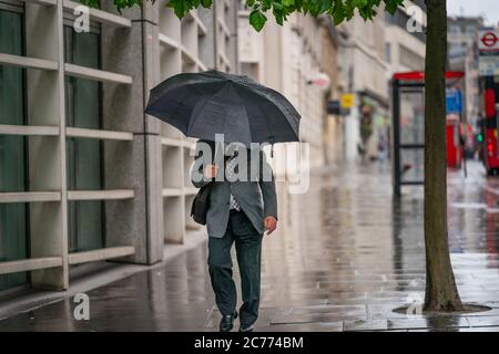 Ein Geschäftsmann mittleren Alters, der einen Anzug trägt, der während eines windigen, nieseligen Tages unter einem Regenschirm in Holborn, London durin, gegen den Wind kämpft Stockfoto