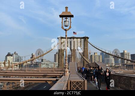 Fußgänger, die von der Brooklyn-Seite der Brooklyn Bridge, New York City, überqueren. Blauer Wolkenhimmel und amerikanische Flagge auf der Brücke. Stockfoto