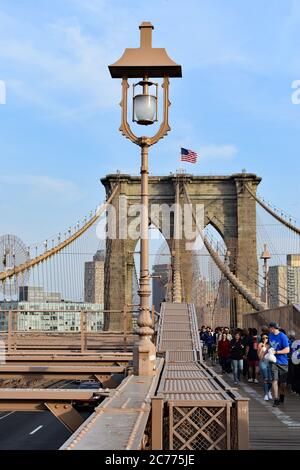 Fußgänger, die von der Brooklyn-Seite der Brooklyn Bridge, New York City, überqueren. Amerikanische Flagge auf der Brücke und altmodische Straßenlaterne. Stockfoto