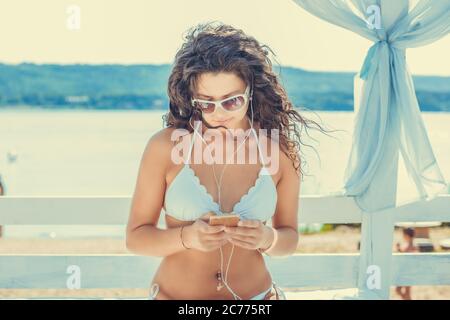 Closeup junge schöne, fröhliche, lächelnde Frau mit Mobil-Handy, die Musik im Freien auf dem Sonnenbett hört, Hintergrund des Seesees sieht aus Stockfoto