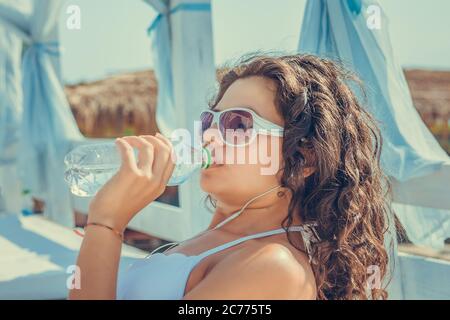 Im Sommer können Sie sich im Freien mit Feuchtigkeit versorgen. Gut aussehende Mixed Race Frau in Sonnenbrille mit Afro Haarschnitt in weißen Badeanzug, Sonne genießen, dri Stockfoto