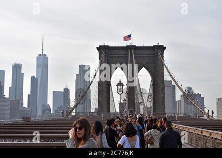 Fußgänger, die die Brooklyn Bridge in New York City überqueren. Der Turm ist unter der amerikanischen Flagge und die Wolkenkratzer der Innenstadt sind hinter gezeigt. Stockfoto