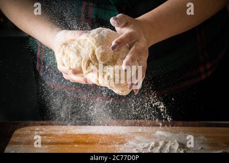 Die Köchin knetet Pizzateig auf einem Holzbrett. Mehl fliegt auf dunklem Grund Stockfoto