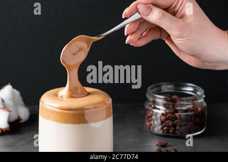 Dalgona Kaffee. Trendiges Getränk auf schwarzem Hintergrund. Frau legt flauschigen süßen Schaum in ein Glas mit Eismilch. Stockfoto