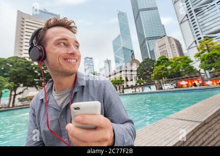 Glücklich junge Kaukasischen Mann Musik auf dem Smartphone hält Handy mit Kopfhörern in der Stadt im Freien Innenstadt von Hongkong, China. Asien Stockfoto