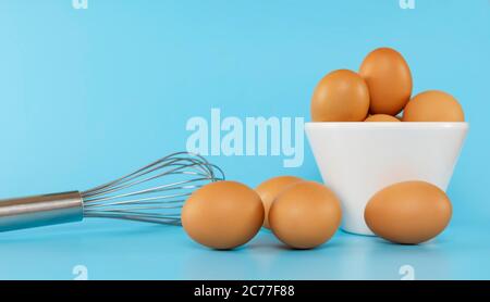 Frische Hühnereier in weißer Schüssel und Hühnereier auf blauem Hintergrund mit Kopierplatz. Rohe Eier in Schüssel und Schneebesen isoliert. Stockfoto