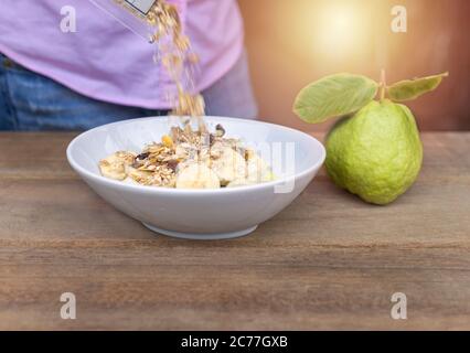 Frauen gießen Haferflocken aus Plastiktüte zu Schüssel und in Schüssel haben Bananenrutsche, Guava auf Holztisch Stockfoto
