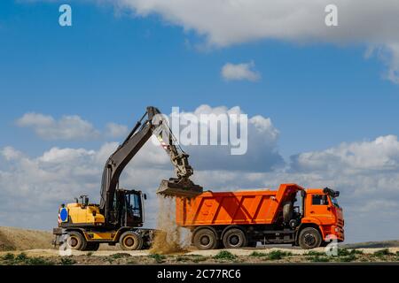 Ein gelber Baggerlader lädt die Erde während des Baus einer Straße gegen einen blauen Himmel mit Wolken in einen LKW. Stockfoto