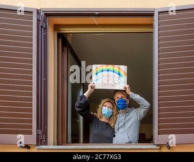 Ein Paar mittleren Alters schaut mit Schutzmasken aus dem Fenster und zeigt das Schild 'Andra' tutto Bene', was auf italienisch bedeutet, dass alles wird Stockfoto