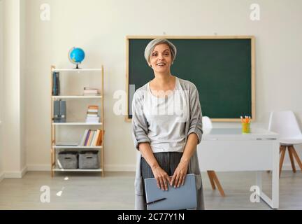 Der Lehrer im Klassenzimmer. Eine Frau mit grauen Haaren eine Lehrerin mit einem Notizbuch in der Hand, das auf dem Hintergrund der Klasse steht. Stockfoto