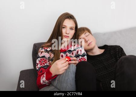 Portrait von schönen Mädchen auf dem Sofa sitzen und traumhaft in der Kamera mit Weihnachtsspielzeug in der Hand suchen, während junge auf sie lehnt und schlafen Stockfoto