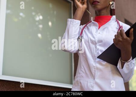 Schwarze junge Ärztin, in einem weißen Mantel, mit einem Phonendoskop, hält eine Mappe für medizinische Papiere in einer Rue, vor dem Hintergrund eines Krankenhauses. Stockfoto