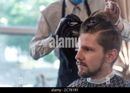 Ein Friseur schneidet einen bärtigen jungen Kerl mit einem Haarschneider und kämmt die Haare auf seinem Kopf. Die Arbeit des Meisters im Männerhaarschnitt in einem Friseurladen Stockfoto