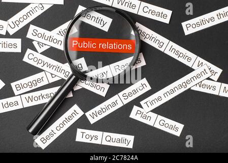 Lupenglas über der roten Aufschrift Public Relations aus Papier geschnitten. Umgeben von anderen Inschriften auf dunklem Hintergrund. Word Cloud-Konzept. Stockfoto
