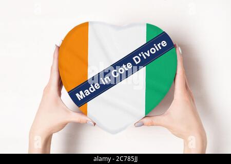 Inschrift hergestellt in der Elfenbeinküste die Flagge der Elfenbeinküste. Weibliche Hände halten eine herzförmige Box. Weißer Hintergrund. Stockfoto