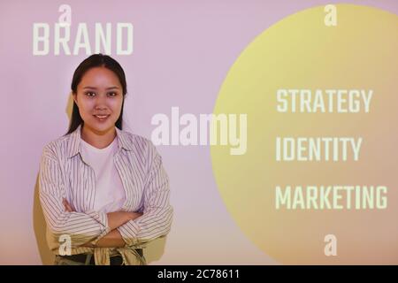 Porträt eines lächelnden, selbstbewussten jungen asiatischen Markenmanagers mit gekreuzten Armen, die gegen die Projektionsleinwand stehen, mit Marketingarbeit Stockfoto