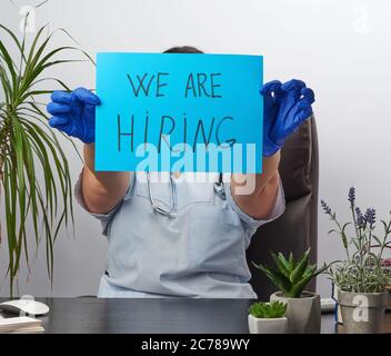 Arzt in einem blauen Mantel, sterile medizinische Handschuhe hält ein Plakat mit der Inschrift wir einstellen, Konzept der Personalausstattung, Mangel an Personal in der Medizin Stockfoto