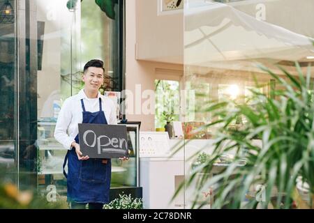 Lächelnder, fröhlicher Cafe-Kellner, der am Eingang steht und ein offenes Schild zeigt, wenn man Gäste begrüßt, nachdem die Sperre beendet ist Stockfoto