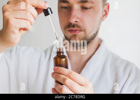 Nahaufnahme von jungen schönen Mann im Bademantel gehen, um Serum oder andere Hautpflege-Produkt auf sein Gesicht gelten. Konzentrieren Sie sich auf die Flasche Stockfoto