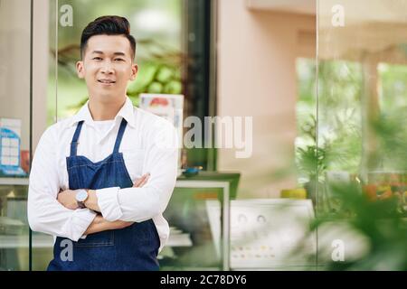 Portrait des selbstbewussten jungen vietnamesischen Café-Besitzers in Denim-Schürze, die mit Armkreuzen vor dem Eingang steht Stockfoto