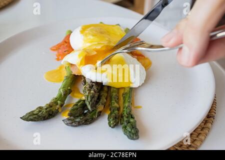 Hände des Mannes schneiden pochiertes Ei mit Sauce Hollandaise und Lachs über Spargel aus der Nähe Stockfoto