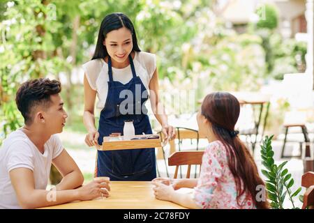 Schöne lächelnde junge asiatische Kellnerin bringt Tablett mit Frühstück zu jungen glücklichen Paar sitzen am Tisch im Café im Freien Stockfoto