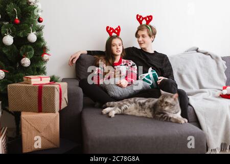 Portrait eines jungen hübschen Paares, das zu Hause auf dem Sofa sitzt und Hirschhörner trägt, während man Zeit mit Katze und Weihnachtsbaum in der Nähe verbringt Stockfoto