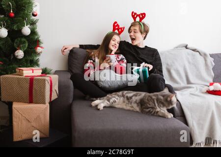 Portrait von jungen schönen Paar sitzt auf dem Sofa zu Hause und trägt Hirsch Hörner, während glücklich Zeit mit Katze in der Nähe verbringen Stockfoto