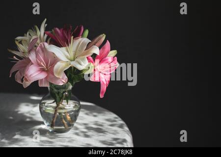 Romantisches Bouquet der weißen und rosa Lilien in einer Glasvase auf dunklem Hintergrund im Sonnenlicht. Für Text platzieren. Stockfoto