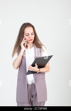 Schöne junge Frau im Geschäft strenge Kleidung stellt sich gegen eine weiße Wand mit einem Telefon in den Händen Stockfoto