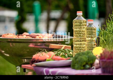 Pflanzenöl auf dem Tisch mit frischem Gemüse auf dem Hintergrund eines Grills im Hinterhof des Hauses. Stockfoto