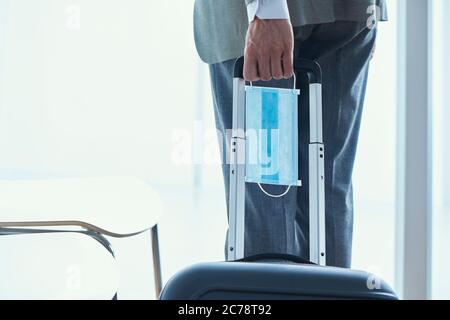 Nahaufnahme eines jungen Geschäftsmannes in einem grauen Anzug, von hinten gesehen, der seinen Koffer am Teleskopgriff zieht und dabei einen chirurgischen Eingriff in der Hand hält Stockfoto