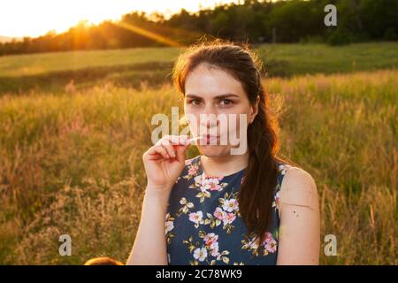 Ein Mädchen in einem gelben Sommerfeld isst Süßigkeiten auf einem Stock und verrunzelt emotional. Abend auf dem Feld und eine Frau. Stockfoto