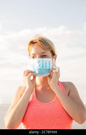 Vertikales Porträt einer schönen blonden Frau trägt eine Schutzmaske mit dem Himmel im Hintergrund, Konzept der Prävention und Gesundheitsversorgung, Kopie sp Stockfoto