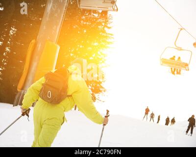 Männliche Skifahrer Skifahren Abfahrt im Skigebiet gegen Skilift. Männlich trägt gelbe Overalls und Rucksack. Nahaufnahme Stockfoto