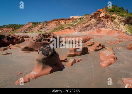 Farbige Steine und Sand am Strand, im Hintergrund Klippen mit natürlicher Vegetation in Praia de Peroba, Icapui, Ceara, Brasilien Stockfoto