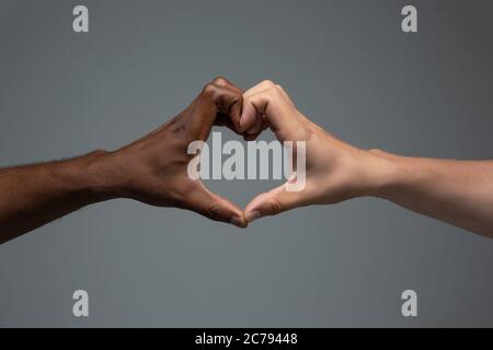 Liebe, Herzensgeste. Rassistische Toleranz. Respektieren Sie die soziale Einheit. Afrikanische und kaukasische Hände gestikulieren auf grauem Studiohintergrund. Menschenrechte, Freundschaft, Konzept der Einheit der Intenationalen. Interrassische Einheit. Stockfoto