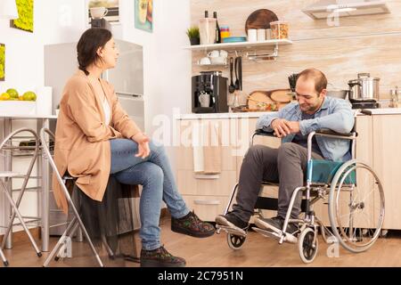 Frau schaut auf behinderten Mann im Rollstuhl, während das Essen auf Gasherd fertig ist. Behinderter gelähmter behinderter Mann mit Gehbehinderung, der nach einem Unfall integriert wird. Stockfoto