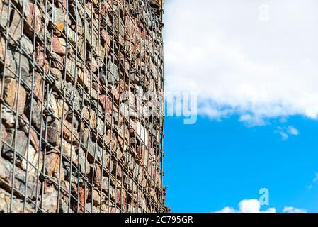 Eine Steinmauer, die an der Straße steht, aus Steinen, die durch Metall geschützt sind, verzinktes Netz im Hintergrund des blauen Himmels mit Wolken, selektiver Fokus. Stockfoto