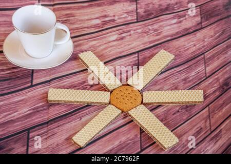 Komposition mit einer Sonne aus Keksen und Waffeln und einer weißen Espressotasse auf einer Tischdecke mit Holzbohlen Stockfoto