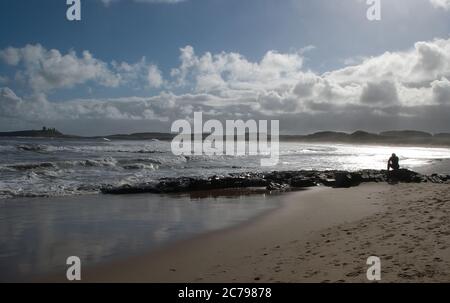 Einzelfigur in Silhouette und tief in Gedanken, während sie auf den Felsen am Wasserrand sitzt und mit Dunstanburgh Castle im Hintergrund Stockfoto